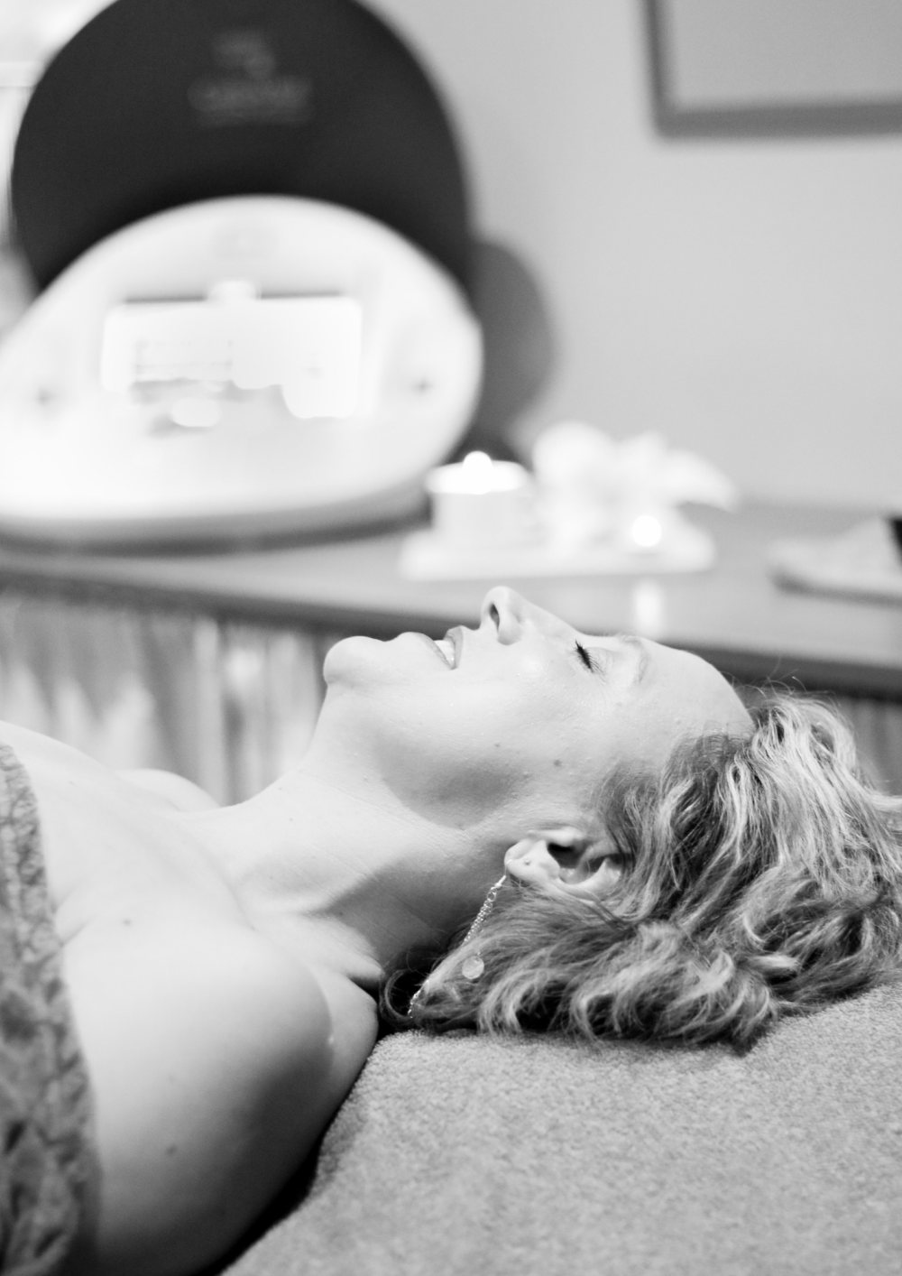 Photo en noir et blanc d'une femme souriante, allongée les yeux fermés sur une table de soins, avec un appareil d'esthétique et une bougie floue en arrière-plan.