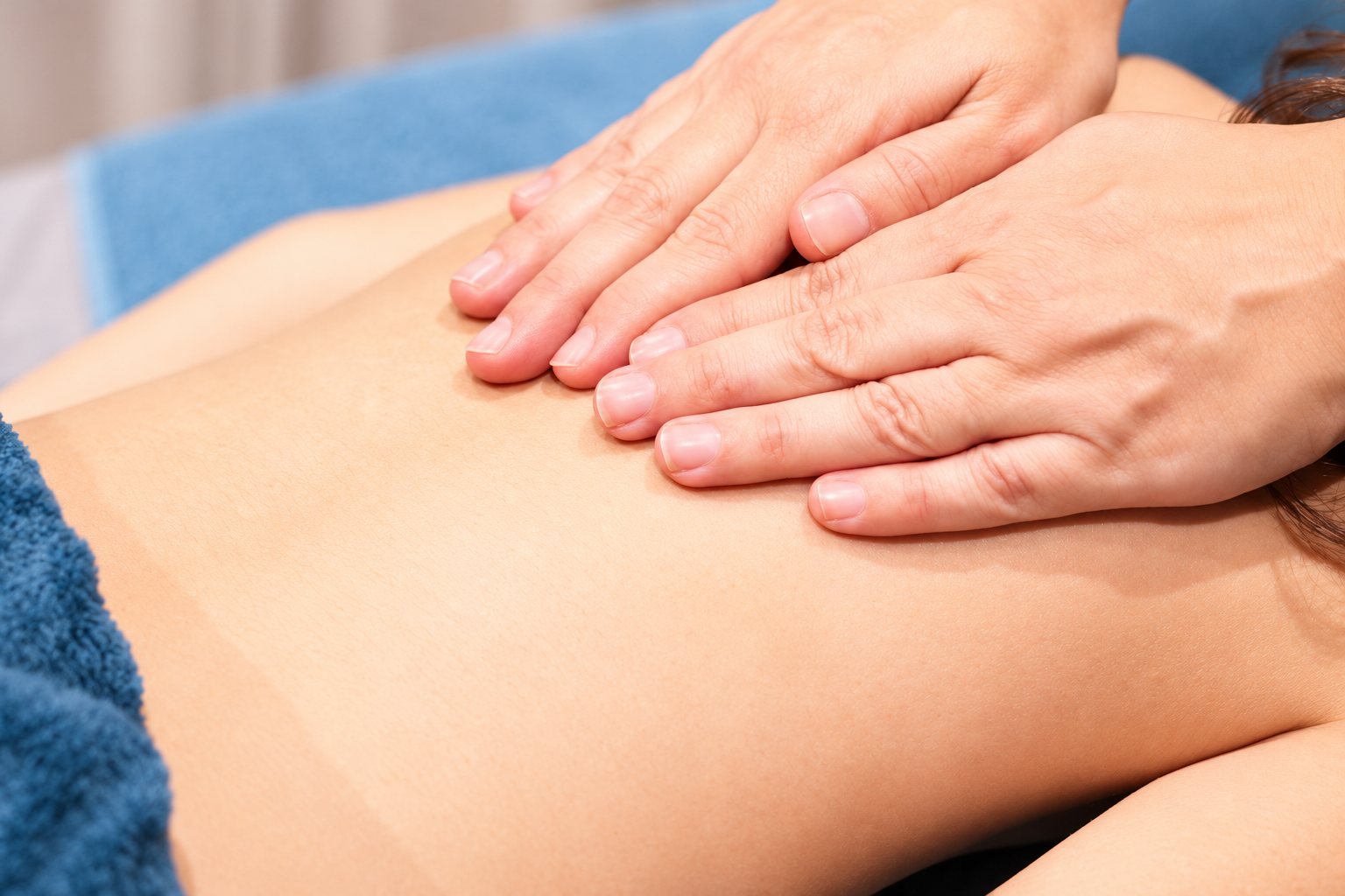Gros plan sur deux mains posées à plat sur le dos nu d'une personne allongée lors d'un massage, avec des serviettes bleues autour.