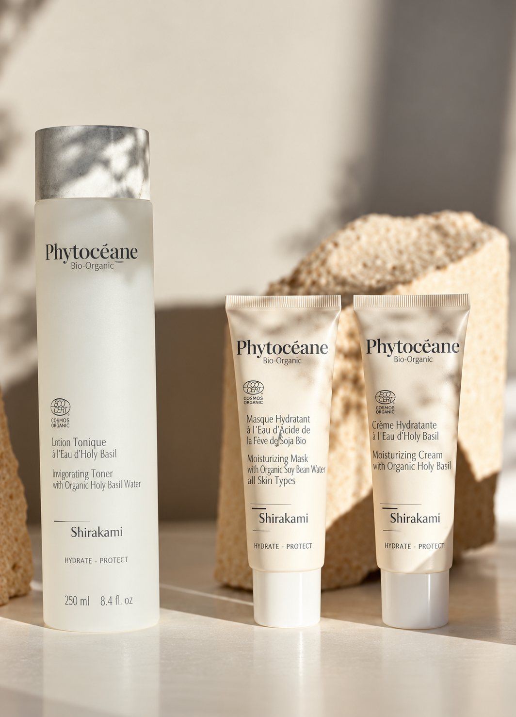 Un flacon et deux tubes de soins cosmétiques Phytocéane de la gamme Shirakami posés sur une surface claire devant une pierre, sous une lumière douce.