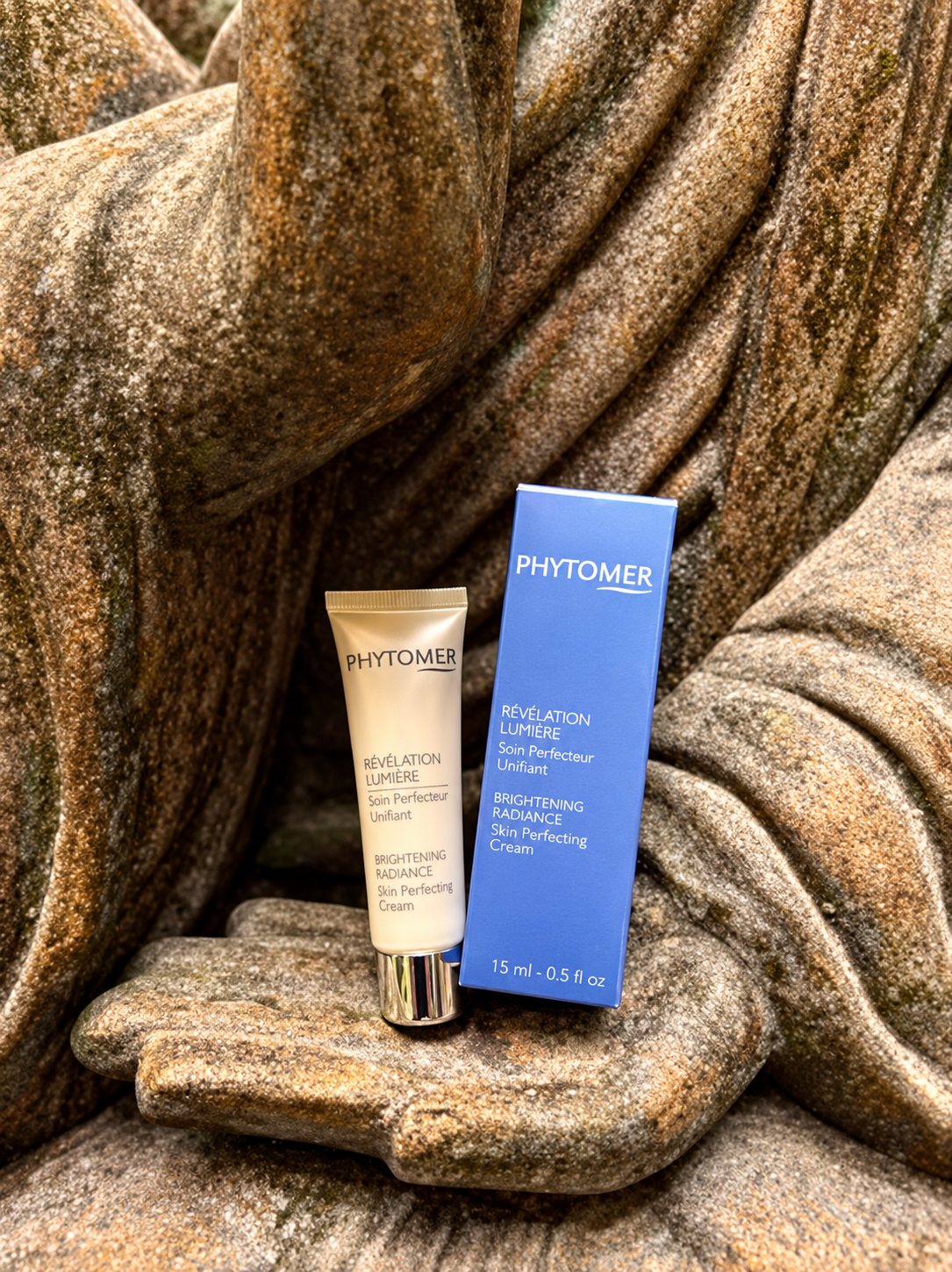 Un tube de crème et sa boîte bleue de la marque Phytomer sont posés dans la main d'une statue de Bouddha en pierre.